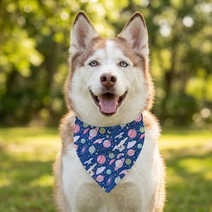 May include: A dog wearing a triangular bandana featuring a dark blue background with a repeating pattern of pink and blue planets, white starships, yellow stars, and white clouds. The dog has light brown and white fur and an open mouth.