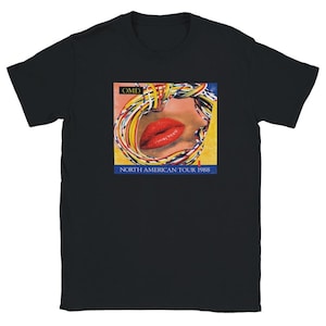 Może przedstawiać: Czarny t-shirt z krótkim rękawem z nadrukiem graficznym przedstawiającym jaskrawoczerwone usta otoczone kolorowym splotem cienkich przewodów. Tekst OMD znajduje się w czarnej ramce u góry, a NORTH AMERICAN TOUR 1988 jest nadrukowany na dole.