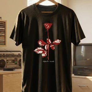 Könnte beinhalten: Dunkelgraues T-Shirt mit einer rot-weißen Rosengrafik. Das Shirt hat den Text "depeche mode" unter der Rose. Das T-Shirt hängt an einem Holzbügel.