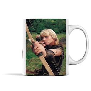 Peut inclure: Une tasse en céramique blanche avec une photo d'un homme tenant un arc et une flèche. L'homme porte une tunique brune et a les cheveux blonds. L'arrière-plan est une forêt verte.