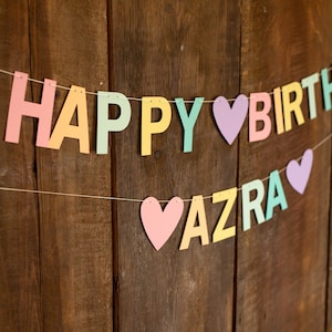 K&ouml;nnte beinhalten: Ein farbenfroher Banner mit den Worten "Happy Birthday Azra" in Pastellfarben mit Herzen zwischen den W&ouml;rtern.
