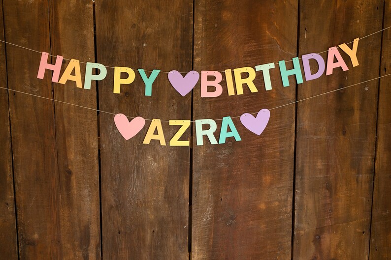 K&ouml;nnte beinhalten: Ein farbenfroher Banner mit den Worten "Happy Birthday Azra" auf einem Holzgrund. Die Buchstaben sind in Pastellfarben gehalten und zwischen den W&ouml;rtern befinden sich Herzen.