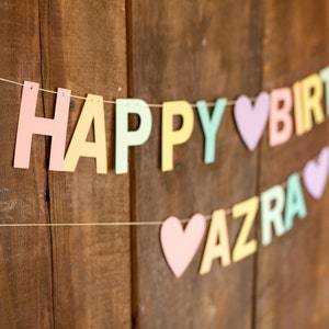 K&ouml;nnte beinhalten: Ein pastellfarbener Banner mit den Worten "Happy Birthday Azra" auf einer wei&szlig;en Schnur vor einem Holz Hintergrund.