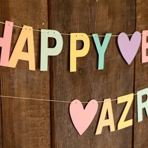 K&ouml;nnte beinhalten: Ein farbenfroher Papierbanner mit den Worten "Happy Birthday Azra" in Pastellfarben. Der Banner ist mit wei&szlig;em Garn aufgef&auml;delt und hat Herzen zwischen den W&ouml;rtern.