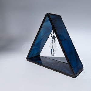 May include: A triangular stained glass art piece with a dark blue hue and a clear crystal pendant hanging from the top. The glass has a marbled effect, and the frame is black. The piece is set against a white background.