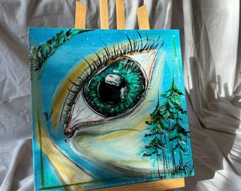 May include: A painting on canvas of a large green eye with a forest scene reflected in the pupil. The eye is painted in a realistic style with detailed eyelashes and a white sclera. The forest scene is painted in a more abstract style with green and blue trees. The painting is on a wooden easel.