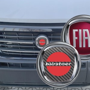 Peut inclure: Gros plan sur l'avant d'un véhicule, avec une calandre chromée et le logo Fiat. Deux emblèmes circulaires avec le logo "bürstner" sont également visibles, l'un sur la calandre et l'autre au premier plan. Le logo Fiat est rouge sur fond bordeaux.