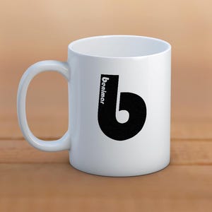May include: White ceramic mug with a large black letter "b" design and the word "benimar" printed vertically. The mug has a curved handle and is set against a wooden surface.