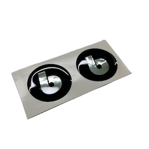 May include: Two black and silver circular stickers with a glossy finish. Each sticker features a silver letter "b" on a black background. The stickers are on a rectangular silver backing.
