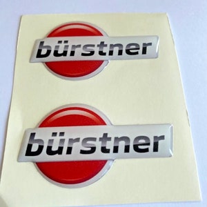 Peut inclure: Deux autocollants blancs avec un emblème 3D rouge et argent. L'emblème porte le mot "bürstner" en lettres noires.