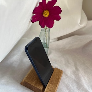 Könnte beinhalten: Ein Handy-Ständer aus Holz mit einem blauen Handy. Der Ständer ist aus hellbraunem Holz gefertigt und hat ein schlichtes, modernes Design. Eine dunkelrosa Blume mit gelber Mitte steht in einer kleinen Glasflasche im Hintergrund.