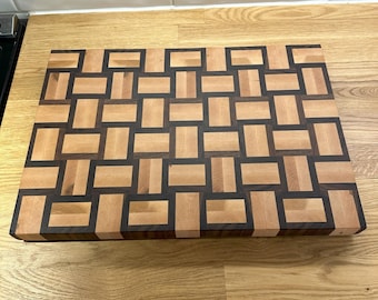 May include: A wooden cutting board with a woven pattern made from light and dark wood.