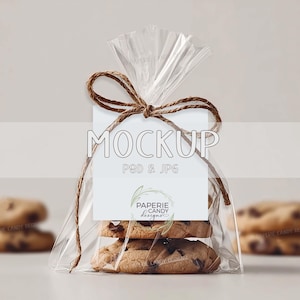 Pode incluir: Uma embalagem transparente de celofane amarrada com barbante, contendo biscoitos com gotas de chocolate. Uma etiqueta branca com a palavra "MOCKUP" e "PSD & JPG" está anexada. A etiqueta também apresenta o logotipo "PAPERIE CANDY designs".