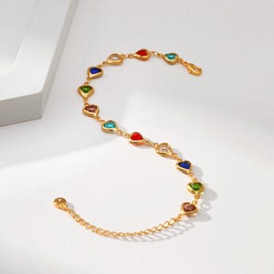May include: Gold-toned bracelet featuring heart-shaped gemstones in various colors, including red, blue, green, and clear. The bracelet has a delicate chain and a secure clasp, perfect for adding a touch of color to any outfit.