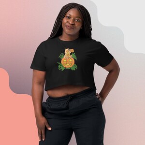 Könnte beinhalten: Ein schwarzes, bauchfreies T-Shirt mit einem Aufdruck eines Comic-Bären, der in einem Kürbis-Gesicht sitzt. Der Bär hält einen kleinen Kürbis. Der Aufdruck ist von grünen Ranken umgeben.