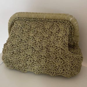 Puede incluir: Bolso de mano tejido en verde claro con un patrón calado texturizado. El bolso tiene una forma redondeada y estructurada con cierre de broche. El material parece ser una fibra natural, lo que le da una estética veraniega e informal.