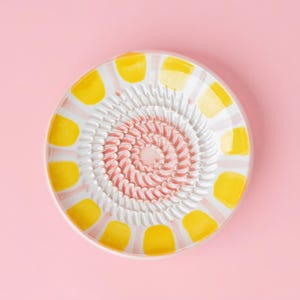 Könnte beinhalten: Ein Keramikreibe mit weißer Mitte und einer rosa spiralförmigen Reibefläche. Der Rand hat gelbe und weiße Farbakzente. Die Reibe befindet sich vor einem rosa Hintergrund.