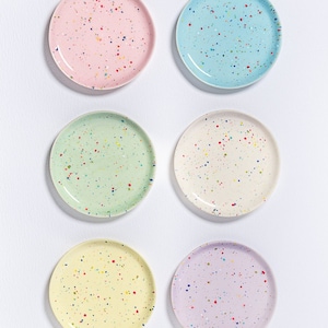 Könnte beinhalten: Sechs gesprenkelte Keramikteller in verschiedenen Pastellfarben, darunter Rosa, Blau, Grün, Gelb und Lila. Die Teller sind in zwei Reihen zu je drei angeordnet, wobei die obere Reihe leicht versetzt zur unteren Reihe ist.