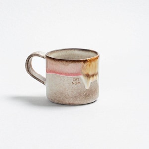Könnte beinhalten: Eine Keramik-Tasse mit Henkel, mit einem zweifarbigen Design aus Rosa und Beige mit braunem Rand. Auf der Tasse steht "CAT MOM". Die Tasse ist für Heißgetränke konzipiert.
