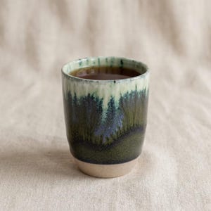 Könnte beinhalten: Ein handgefertigter Keramikbecher mit einer von Wäldern inspirierten Glasur. Der Becher hat einen hellbeigen Boden, der in einen dunkelgrünen und blauen Mittelteil und einen hellgrünen Rand übergeht. Der Becher ist mit einer dunklen Flüssigkeit gefüllt.