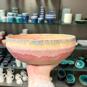 Könnte beinhalten: Eine große, handgefertigte Keramikschale mit einem breiten, flachen Design. Die Schale hat einen Farbverlauf, der von einer hellrosa Basis zu einem gelben und silbernen Rand übergeht. Die Schale wird in einer Hand gehalten, um ihre Größe und Form zu zeigen.