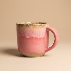 Könnte beinhalten: Ein Keramikbecher mit einer glänzenden rosa Glasur, die zum oberen Rand hin in einen helleren, cremefarbenen Rosaton übergeht. Der Rand selbst weist ein gesprenkeltes braunes und beiges Finish auf. Der Becher hat einen geschwungenen Henkel.