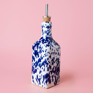 May include: A white ceramic bottle with a blue splatter design and a silver pour spout. The bottle is on a pink background.