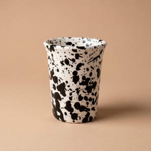 Könnte beinhalten: Ein Keramikbecher mit weißem Untergrund und einem schwarzen Farbspritzermuster. Das Gefäß hat eine zylindrische Form mit einem leicht ausgestellten Rand und einer glatten Oberfläche, die ein abstraktes Design aus schwarzen Flecken zeigt.