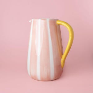 Könnte beinhalten: Eine hohe Keramikkaraffe mit rosa und weißen Streifen und einem gelben Griff. Der Hintergrund ist einfarbig rosa. Diese dekorative Karaffe eignet sich zum Servieren von Getränken. Die Höhe beträgt ca. 25 cm.
