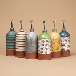 Könnte beinhalten: Sechs Keramik-Ölspender mit Edelstahl-Ausgießern, jeder mit einem eigenen Muster wie horizontalen Streifen, Punkten und geometrischen Formen. Die Flaschen haben einen Terrakotta-Boden und einen glasierten oberen Teil in Blau, Weiß, Grün und Gelb.