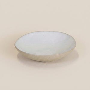 Könnte beinhalten: Eine kleine, cremefarbene Keramikschale mit leicht unregelmäßiger, ovaler Form. Der Innenraum ist ein helles, gesprenkeltes Weiß, das mit dem strukturierten, beigefarbenen Äußeren kontrastiert. Die Schale steht auf einer neutralen Oberfläche.