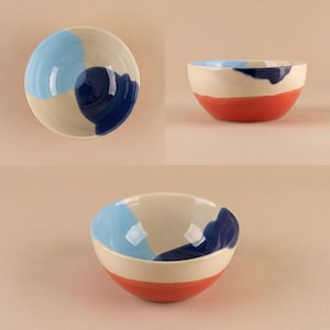 Könnte beinhalten: Eine Keramikschale mit cremefarbener Basis und einem breiten orangefarbenen Band am Boden. Das Innere und die obere Außenseite sind mit abstrakten, handgemalten Spritzern aus hellblauem und dunkelmarineblauem Glasurfinish versehen.