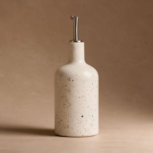 Könnte beinhalten: Eine cremefarbene Keramik-Ölspenderflasche mit einem gesprenkelten Muster aus kleinen Punkten in verschiedenen Farben. Die Flasche hat einen zylindrischen Körper, einen schmalen Hals und einen silberfarbenen Ausgießer aus Metall.