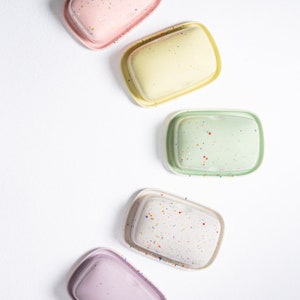Könnte beinhalten: Fünf bunte Keramik-Butterdosen mit gesprenkelten Mustern. Die Dosen sind in einer Reihe angeordnet, wobei die rosa Dose oben und die lila Dose unten ist.