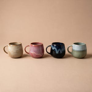 Könnte beinhalten: Vier Keramikbecher in einer Reihe, jeder mit einer einzigartigen zweifarbigen Reaktivglasur. Die Farben umfassen Beige und Creme, Pink und Rosa, Dunkelblau und Schwarz sowie Salbeigrün und Hellblau, alle mit abgerundeten Körpern und Henkeln.