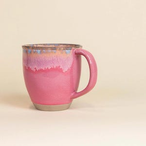 Könnte beinhalten: Eine rosa Keramiktasse mit rundem Körper und gebogenem Henkel. Der obere Rand weist einen Farbverlauf auf, darunter Blau, Rosa und Pfirsich. Der Boden der Tasse ist in einem neutralen Beigeton gehalten. Die Tasse ist für Heißgetränke konzipiert.