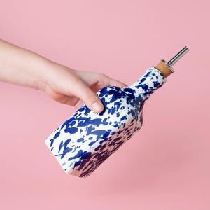 May include: A white ceramic bottle with a blue splatter design and a silver pour spout. The bottle is being held by a hand.