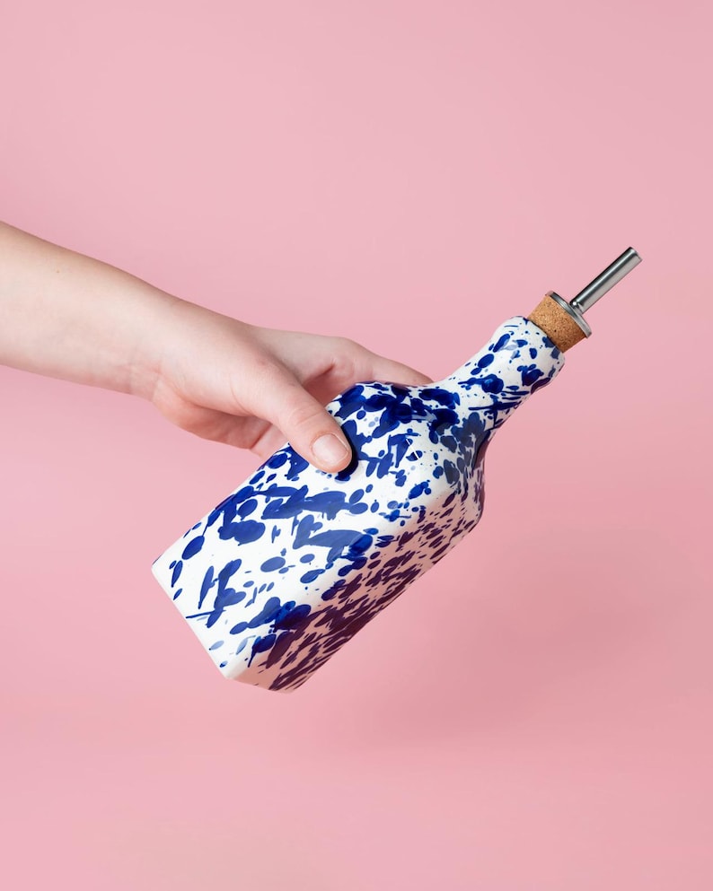 May include: A white ceramic bottle with a blue splatter design and a silver pour spout. The bottle is being held by a hand.