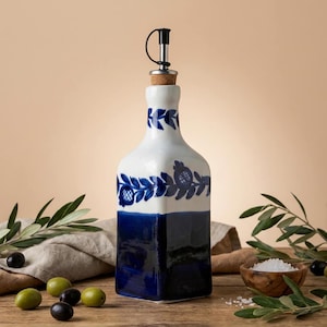 Könnte beinhalten: Eine weiße und blaue Keramikölflasche mit floralem Design. Die Flasche hat einen dunkelblauen Sockel und einen Korken mit Metallschnabel. Oliven und Olivenzweige sind um die Flasche auf einer Holzoberfläche angeordnet.