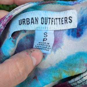 Peut inclure: Un chemisier tie-dye avec une &eacute;tiquette blanche qui dit "Urban Outfitters" et "S P" avec le texte "Made in Cambodia, Fabriqu&eacute; au Cambodge".