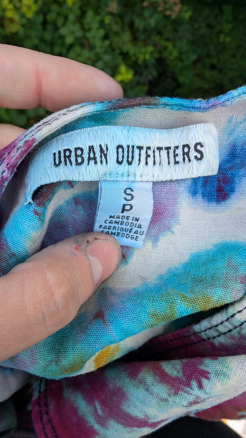 Peut inclure: Un chemisier tie-dye avec une &eacute;tiquette blanche qui dit "Urban Outfitters" et "S P" avec le texte "Made in Cambodia, Fabriqu&eacute; au Cambodge".