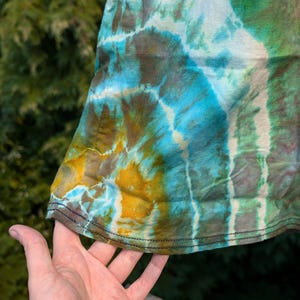 Peut inclure: Un tissu tie-dye en gros plan avec un motif tourbillonnant de vert, bleu, jaune et blanc. Le tissu est pli&eacute; et les bords sont cousus.