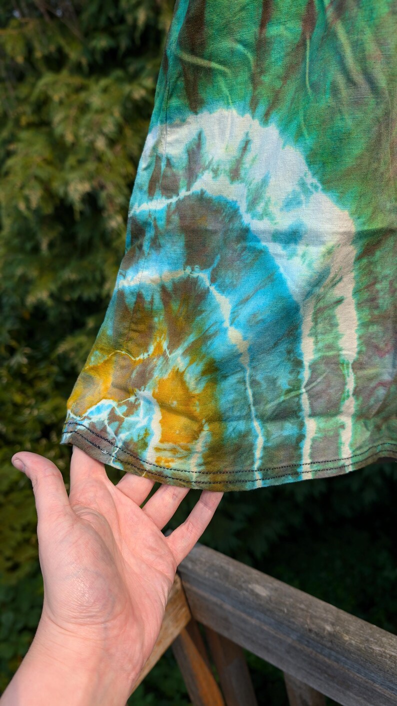 Peut inclure: Un tissu tie-dye en gros plan avec un motif tourbillonnant de vert, bleu, jaune et blanc. Le tissu est pli&eacute; et les bords sont cousus.