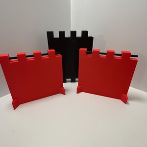 May include: Three-dimensional, geometric castle wall pieces in red and black. Two red pieces are in the foreground, with a black piece behind them. Each piece has a rectangular shape with a crenelated top and a small base.