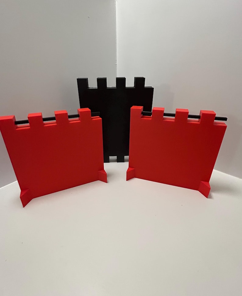 May include: Three-dimensional, geometric castle wall pieces in red and black. Two red pieces are in the foreground, with a black piece behind them. Each piece has a rectangular shape with a crenelated top and a small base.