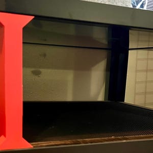 May include: A black and red terrarium stand with a glass enclosure. The stand features a red support structure with a black base and top. The enclosure has a black frame and a mesh bottom.