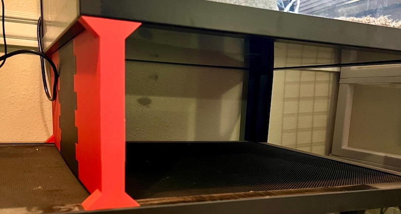 May include: A black and red terrarium stand with a glass enclosure. The stand features a red support structure with a black base and top. The enclosure has a black frame and a mesh bottom.