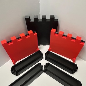 May include: A set of black and red castle-themed building blocks. The set includes three red crenelated wall pieces, one black crenelated wall piece, and four black base pieces. The blocks are arranged on a white surface.