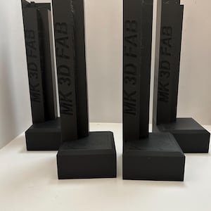 Puede incluir: Cuatro objetos negros verticales en forma de L con bases rectangulares. Cada pieza presenta el texto MK 3D FAB grabado verticalmente en el lateral. Los elementos están dispuestos en dos pares, de pie con un acabado mate y bordes geométricos.