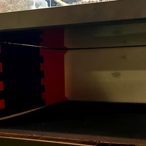 May include: A black and red terrarium with a black wire and a mesh bottom. The terrarium has a red support structure with a black, interlocking design. The background is a light beige color.